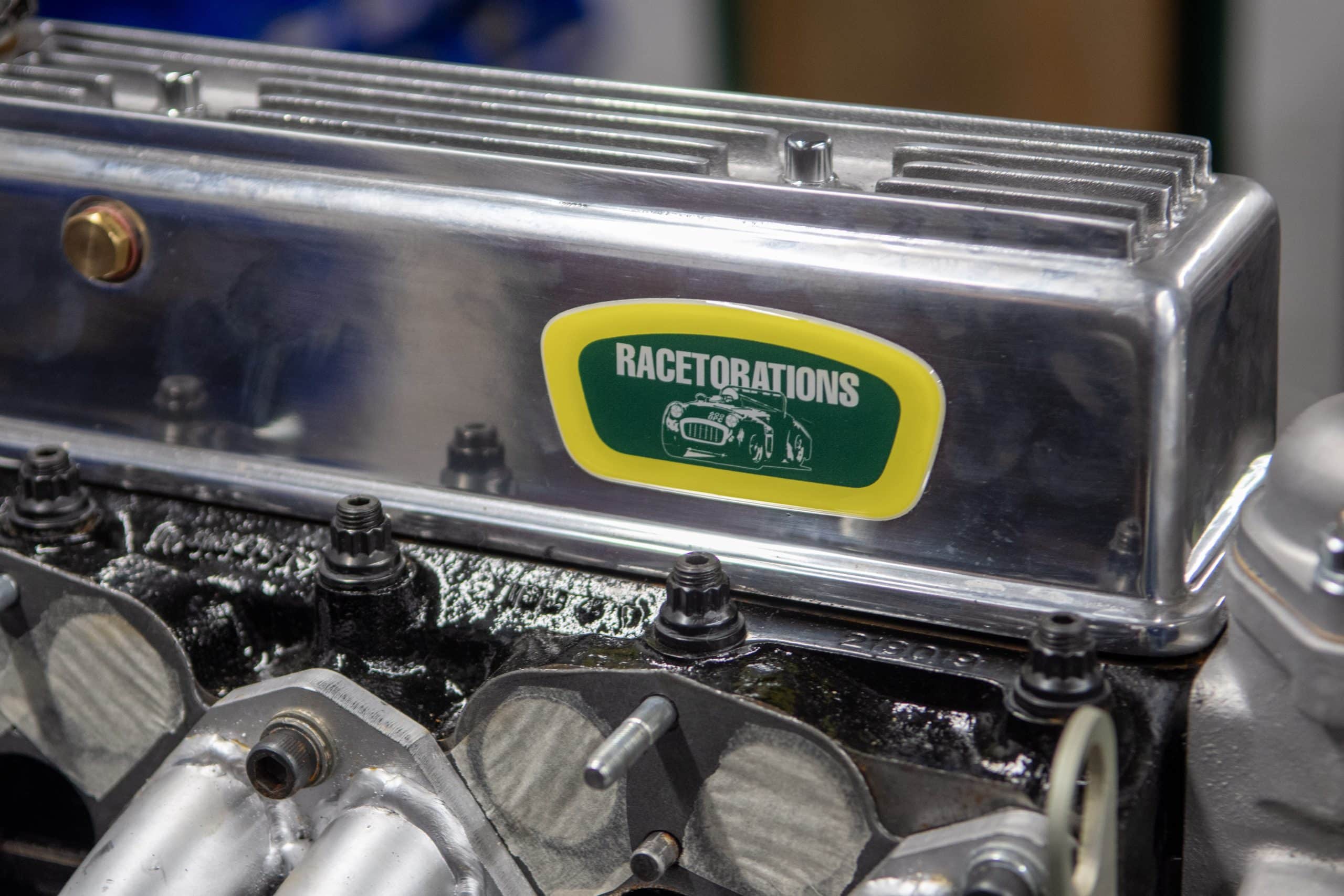 Close-up of a car engine part with a silver cover. A yellow and green label reads "RACETORATIONS," featuring a silhouette of a race car. The engine has several bolts and a metallic finish.