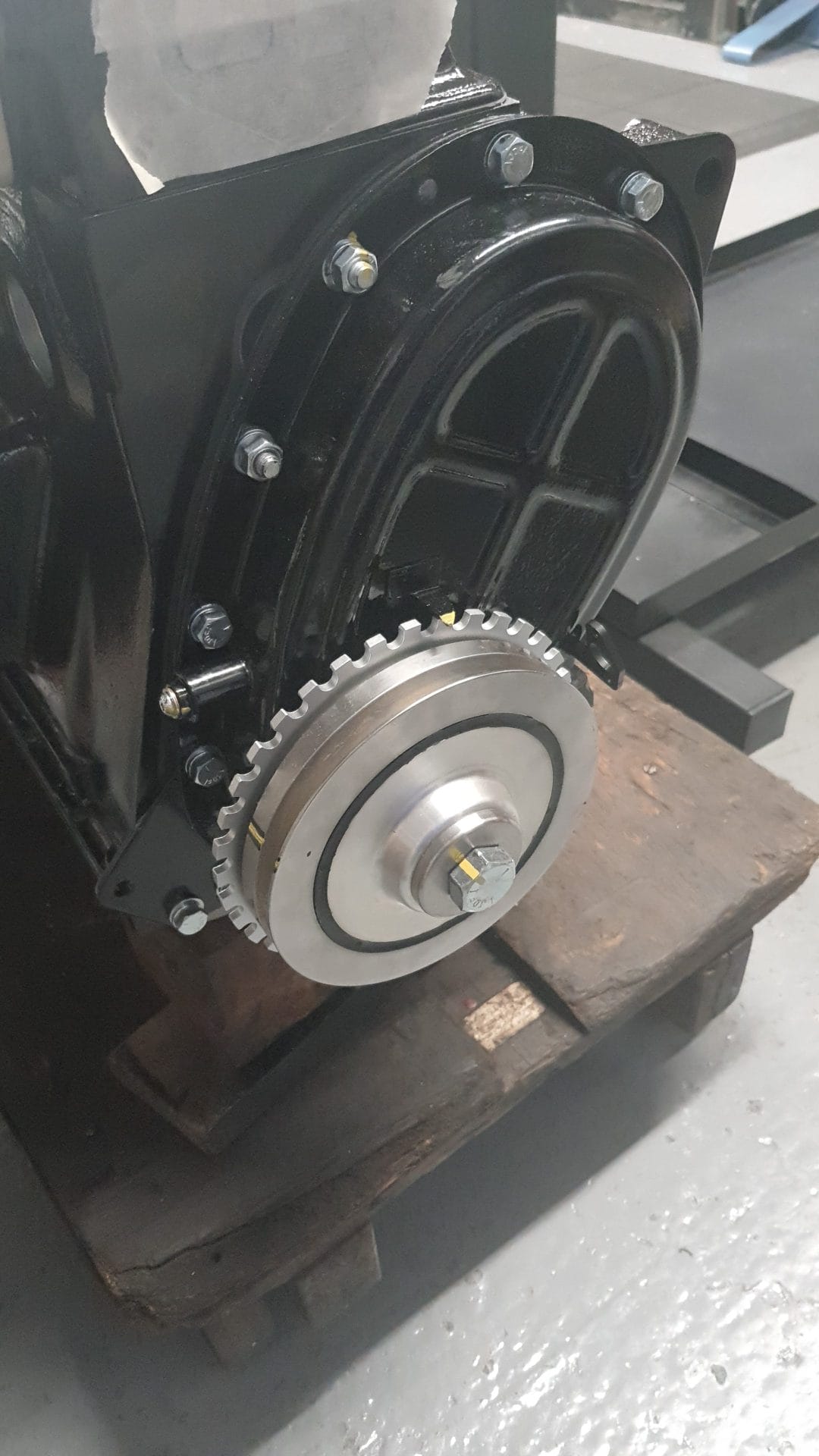Close-up of the Racetorations Crankshaft Damper Trigger Wheel and pulley assembly fitted to a black engine block, all mounted on a wooden pallet on a grey industrial floor.