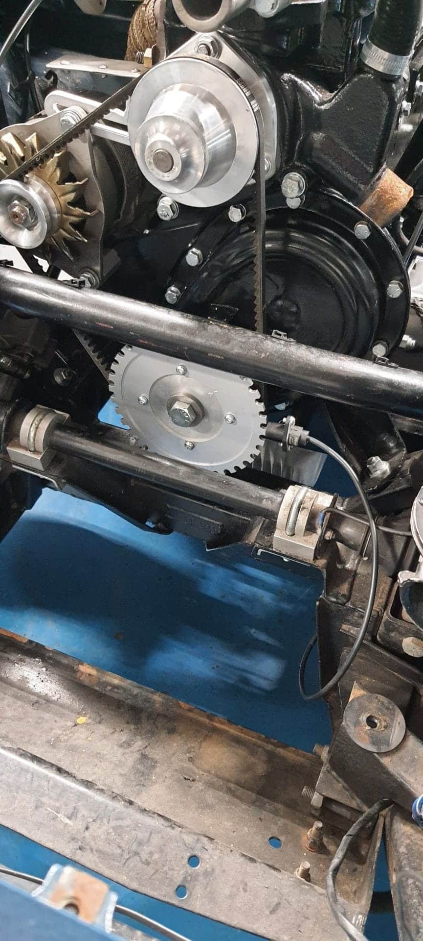 Close-up of a car engine with pulleys and gears, featuring the Racetorations Crankshaft Damper Trigger Wheel fitted to metallic parts, all securely mounted on a black Racetorations frame on a blue floor.