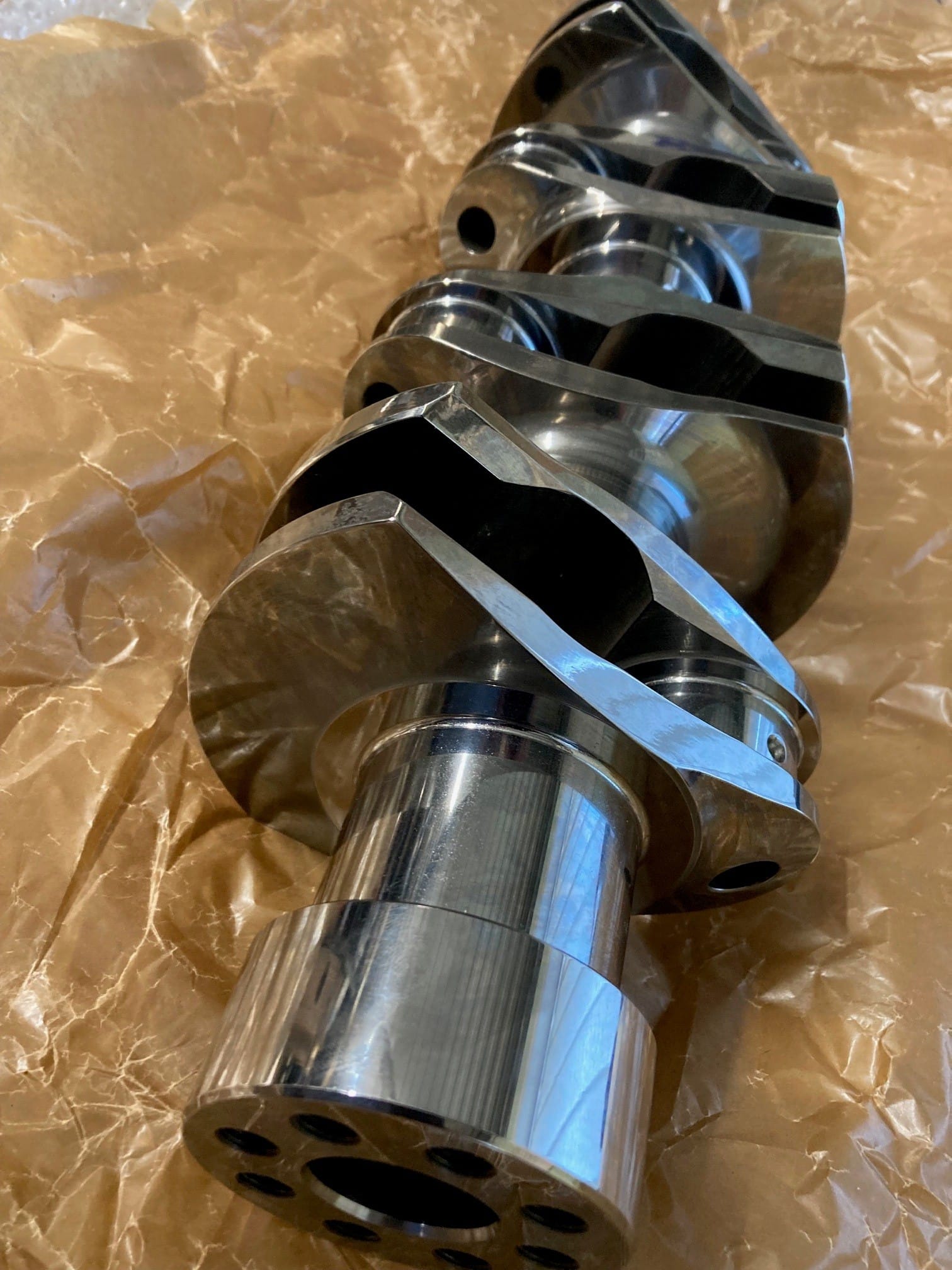 The Arrow Precision Engineering Billet Steel Crankshaft, Narrow Journal - TR2-4, Morgan +4, expertly crafted through precision engineering, rests on crumpled brown paper, its machined surfaces gleaming in the light.