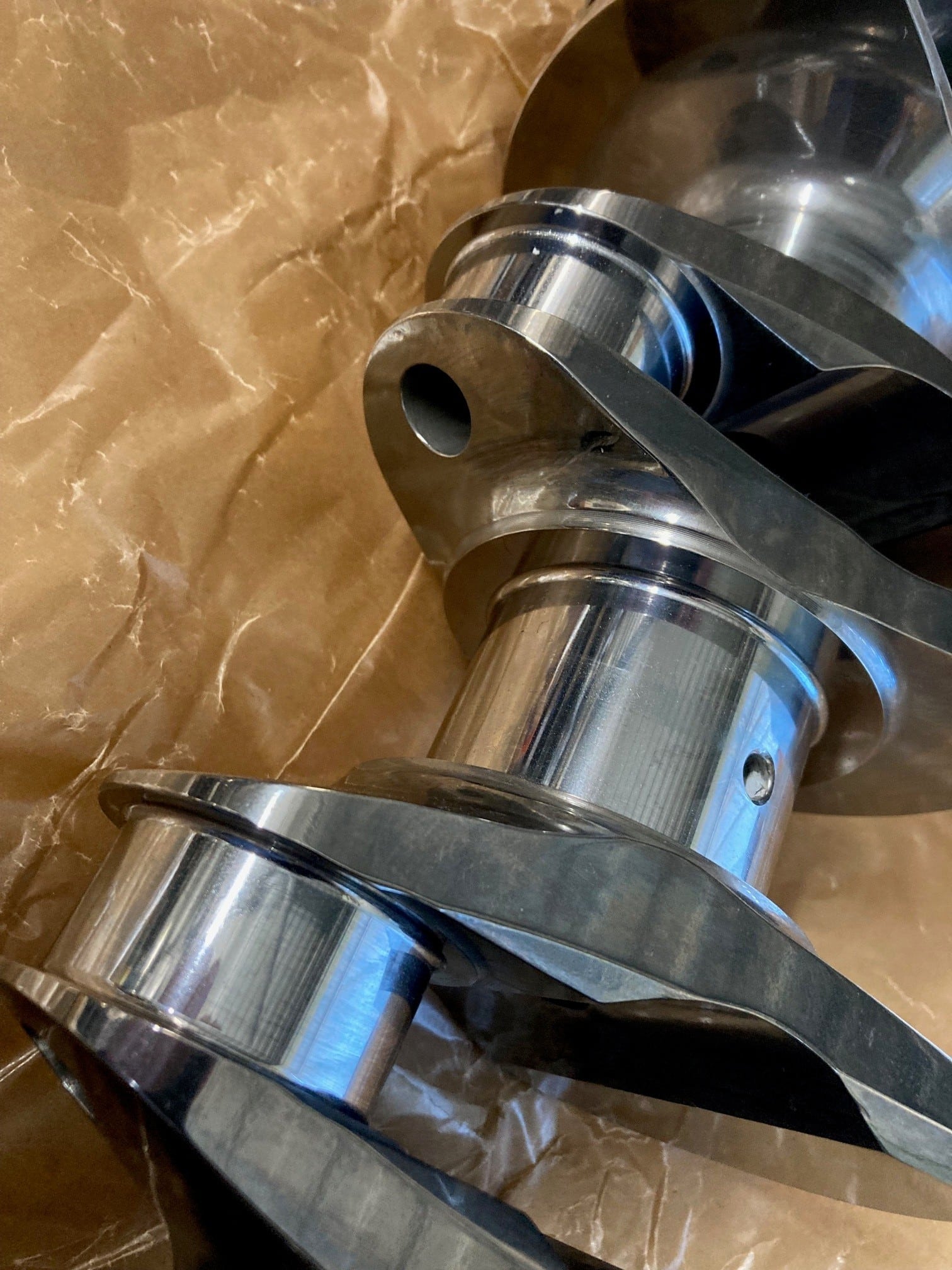 A close-up of a shiny Arrow Precision Engineering Billet Steel Crankshaft, Narrow Journal – TR2-4, Morgan +4, resting on crumpled brown paper, highlighting its smooth surfaces and precise machined details—an expertly crafted component engineered for performance and reliability.