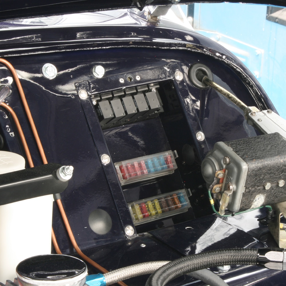 Close-up of the Racetorations Hidden Fuse & Relay Box – TR2-3A, mounted in the engine bay. Fuses and relays are neatly arranged with wiring amid metallic parts, reflecting light beneath the open bonnet.