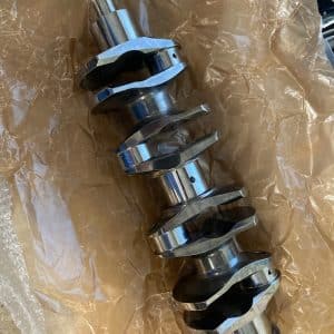 A billet steel crankshaft resting on brown packing paper and bubble wrap, viewed from above. The crankshaft features multiple machined lobes, narrow journal bearing surfaces, and a superfinished appearance, indicating it is new or recently manufactured.