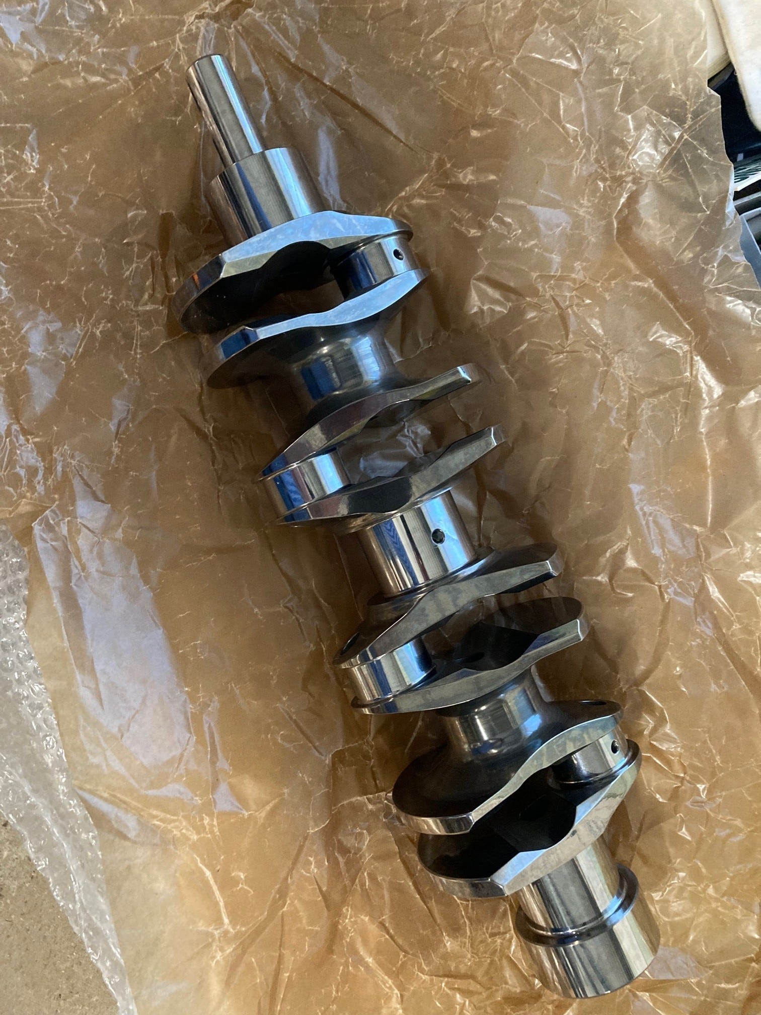 A billet steel crankshaft resting on brown packing paper and bubble wrap, viewed from above. The crankshaft features multiple machined lobes, narrow journal bearing surfaces, and a superfinished appearance, indicating it is new or recently manufactured.