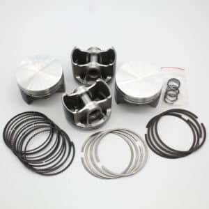 Four metal pistons and various piston rings are arranged on a white surface. The rings are grouped in stacks and some are separated, whilst a small plastic bag contains additional small rings.
