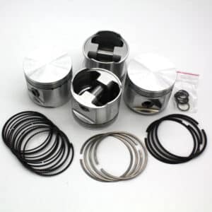 Four metal engine pistons are arranged on a white surface, along with several black and silver piston rings and a small plastic bag containing additional small parts.