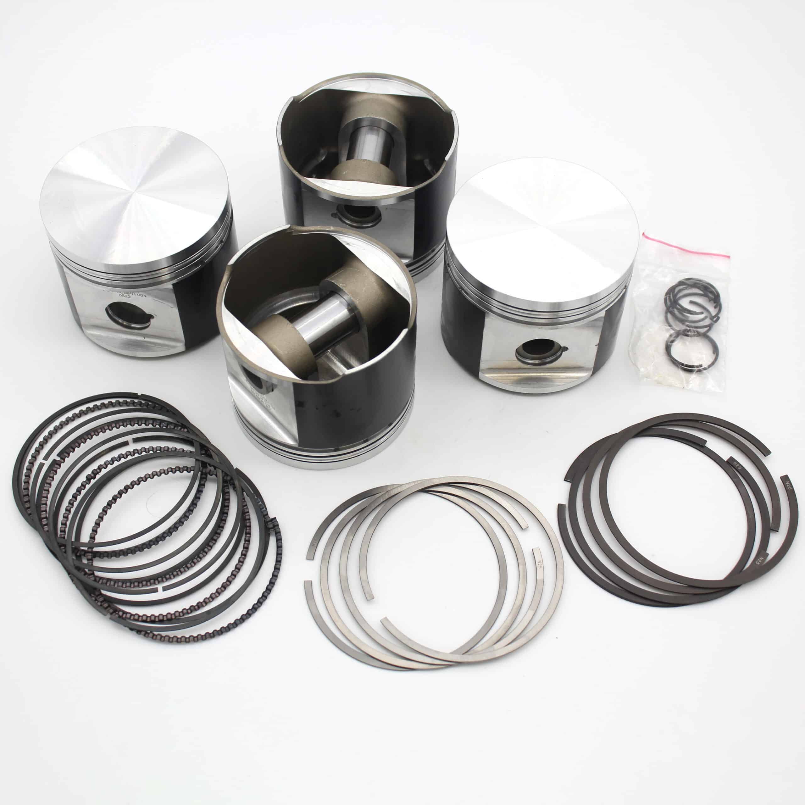 Four metallic engine pistons with connecting rods are arranged on a white surface, along with several sets of piston rings and a small plastic bag containing additional rings or clips.