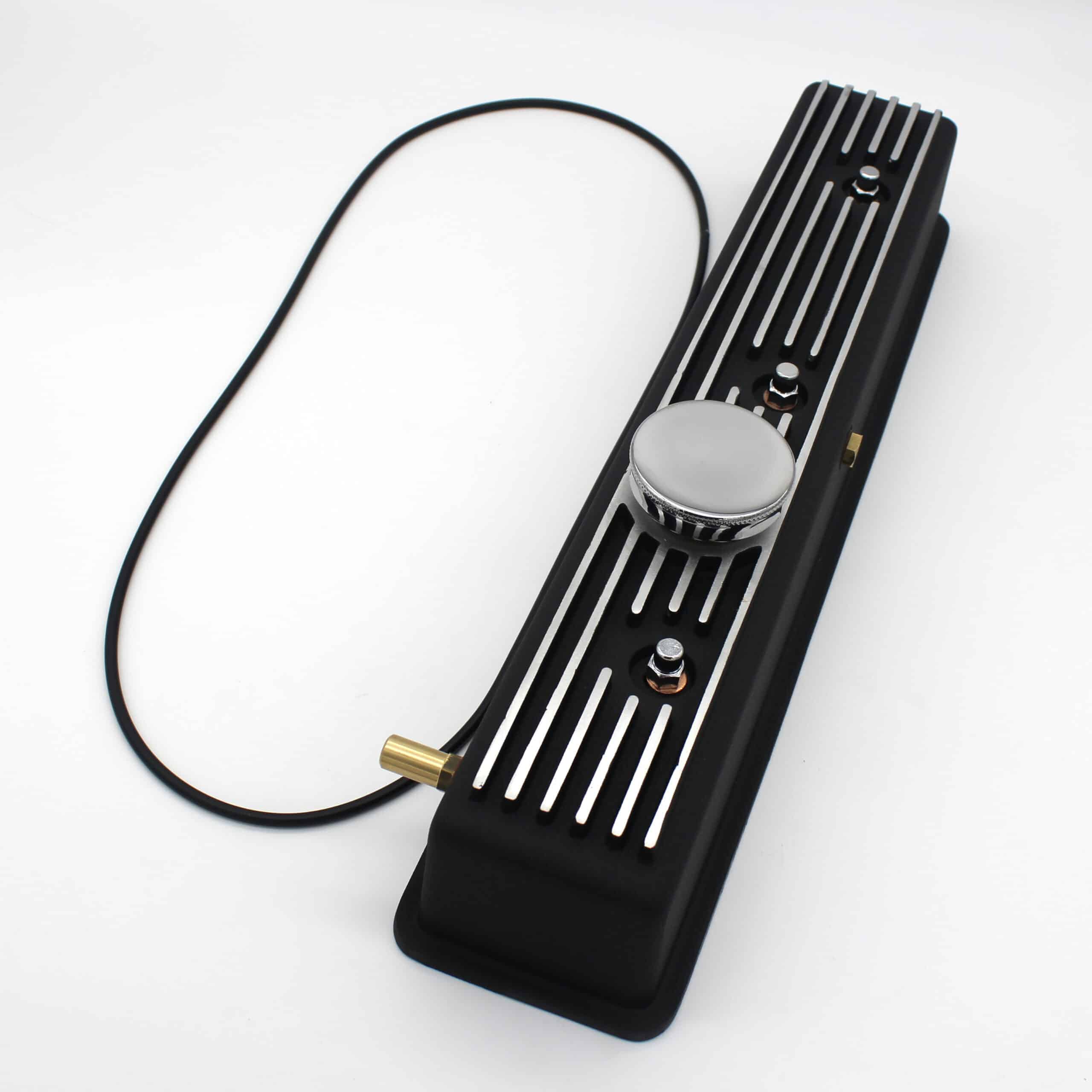 A black, rectangular electronic device with metallic stripes on top, a large round knob in the centre, smaller knobs, and a connected cable, set against a white background.