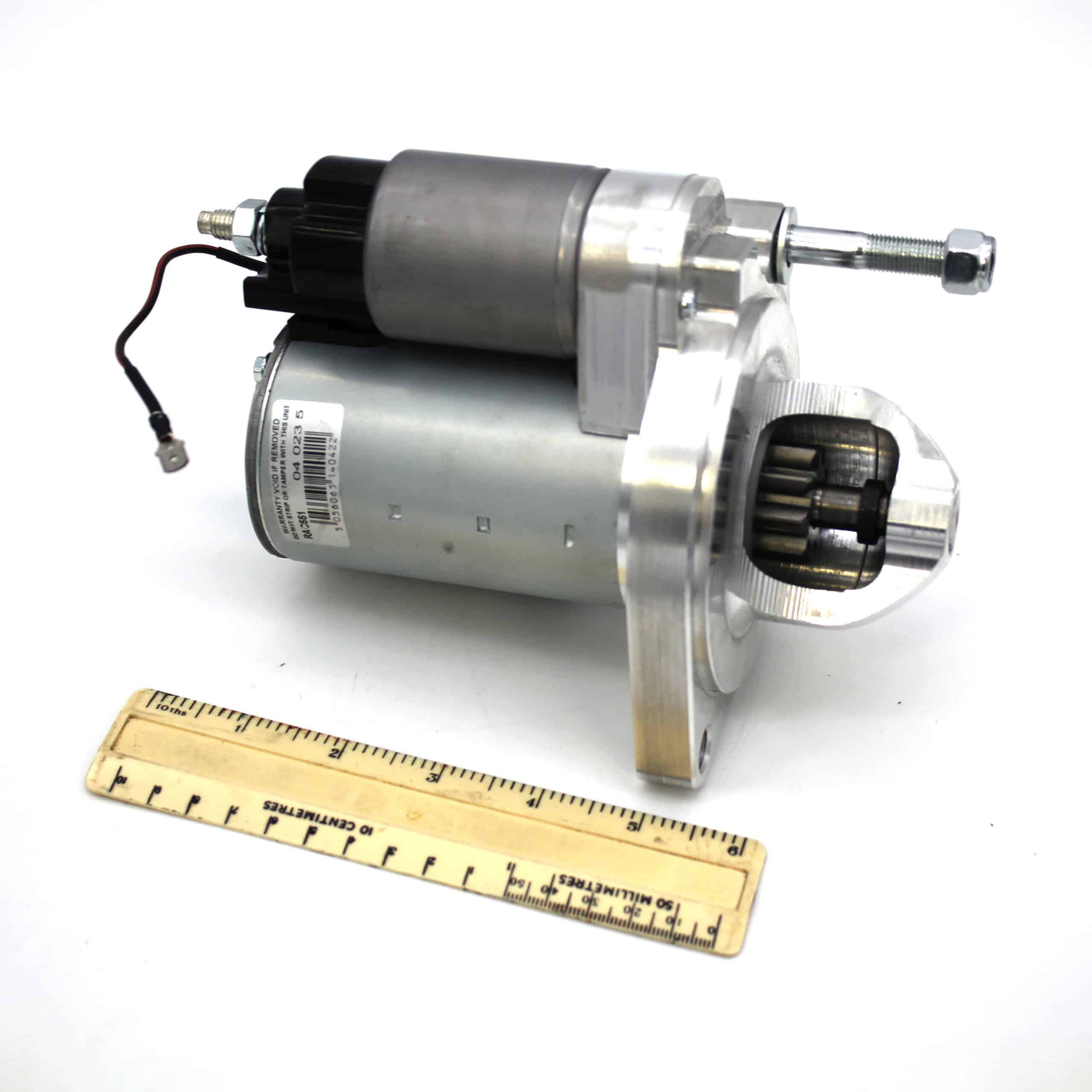 A metallic starter motor with visible gears is placed next to a wooden ruler for scale, on a white background. A wire extends from the motor, and the ruler measures approximately 30 centimetres.