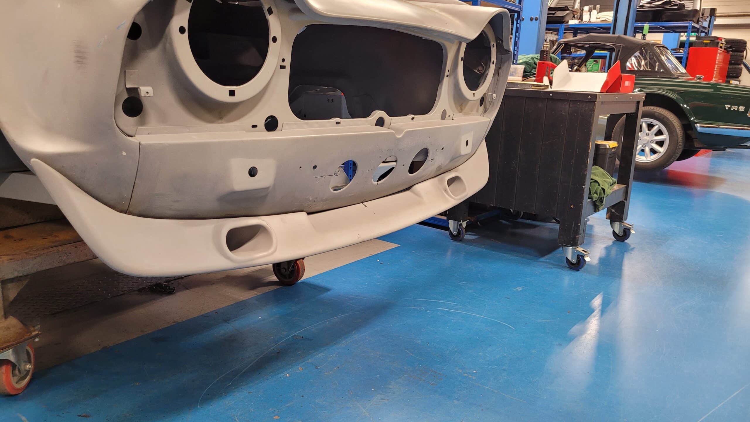 Close-up of a Triumph TR4 with its front end stripped, highlighting the Racetorations Front Spoiler With Brake Cooling Ducts. The car is in a restoration workshop with tools, blue floor, and a green classic car seen in the background.