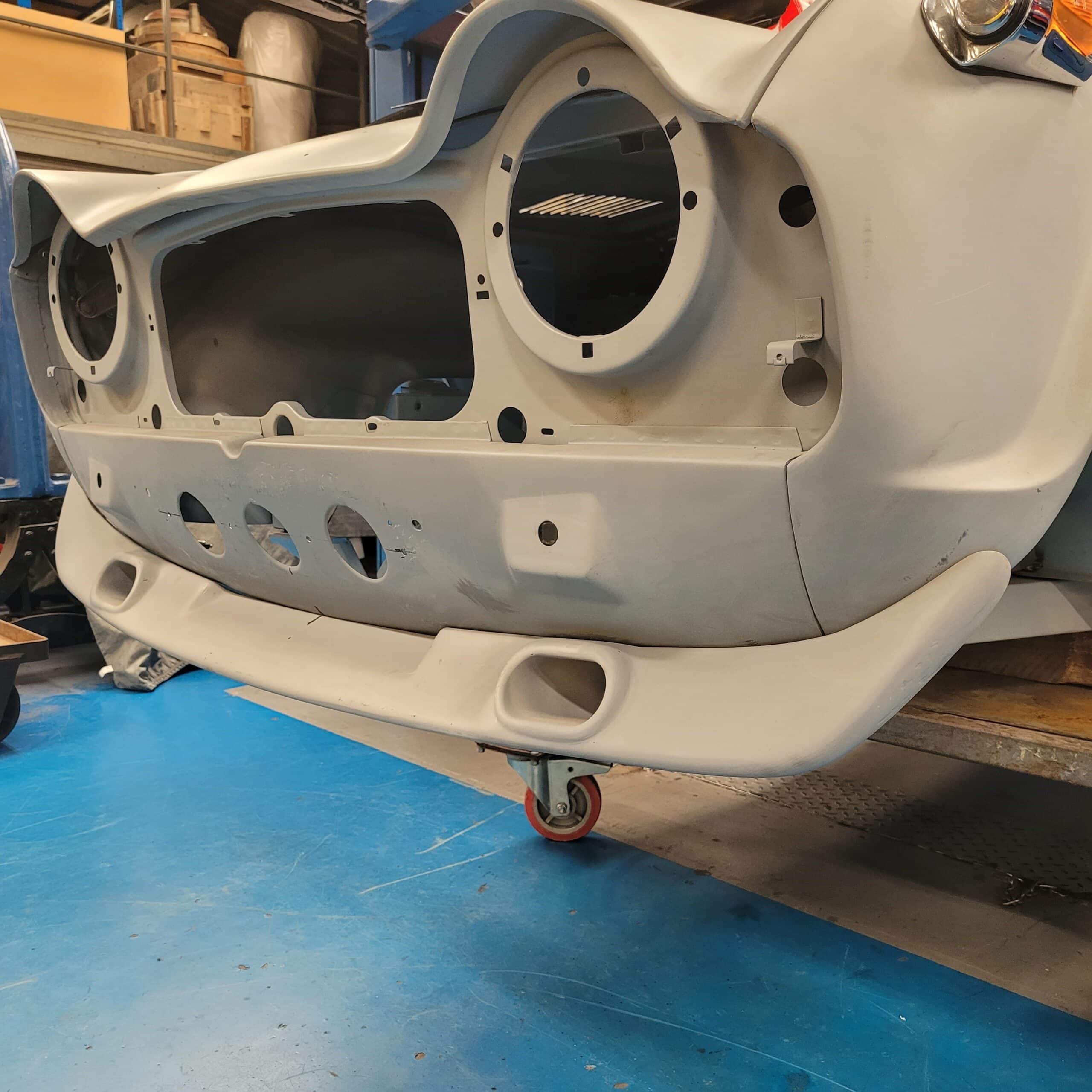 A Racetorations Front Spoiler With Brake Cooling Ducts for Triumph TR4, TR4A, TR5, or TR250 shown unpainted on a blue workshop floor, featuring empty headlamp and grille openings and mounted on small wheels for easy movement.