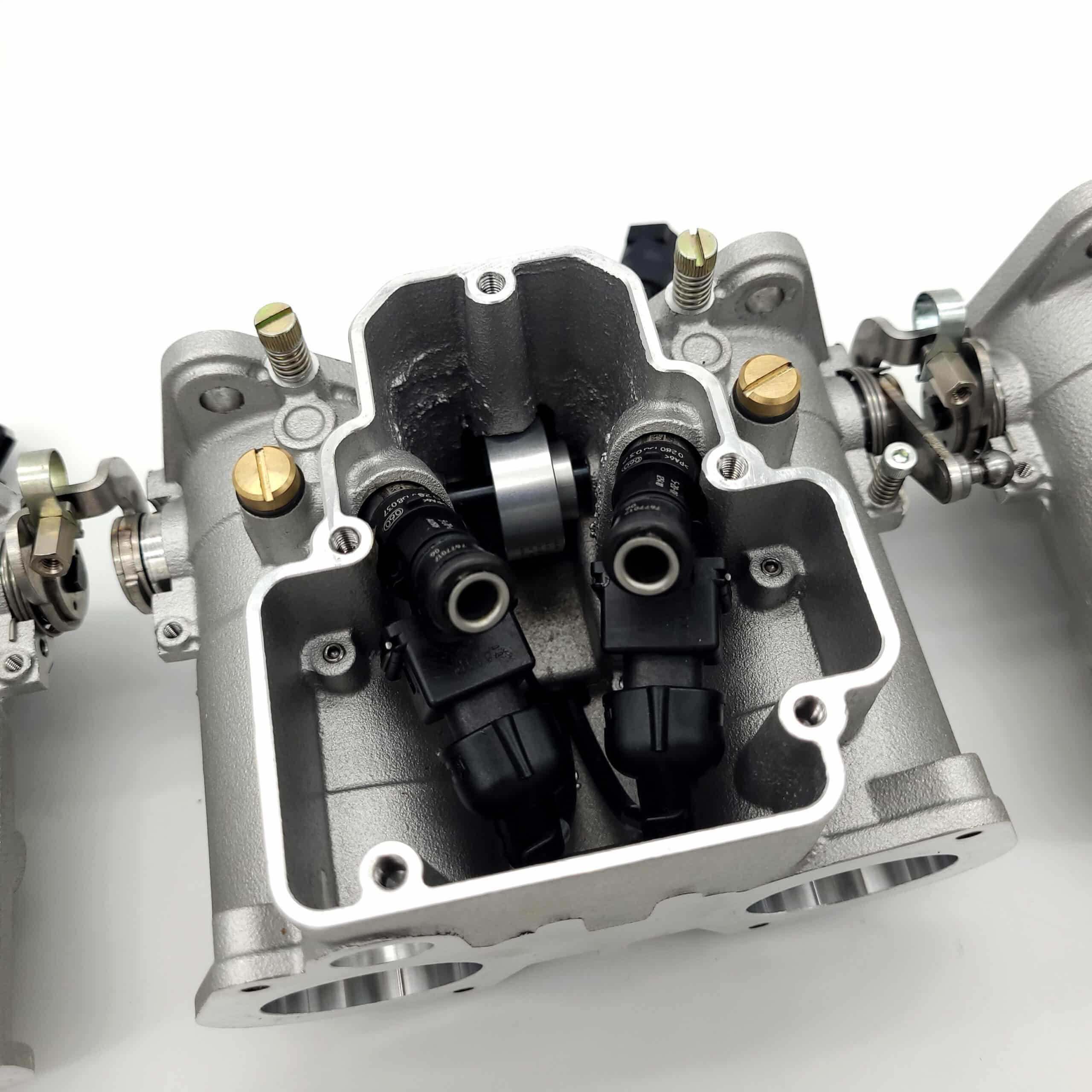 Close-up of a Jenvey Heritage Throttle Body Set - TR5-6, highlighting two fuel injectors and multiple screws, bolts, and cylindrical components against a white background.