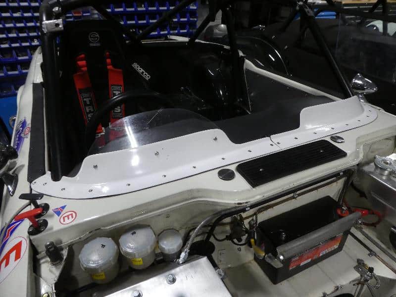 Close-up of a TR6 race car cockpit with racing seat, harness, steering wheel, control panel, fluid reservoirs, and battery. A Racetorations Wind Deflector - TR4-6 is fitted beside visible cables and mechanical components.
