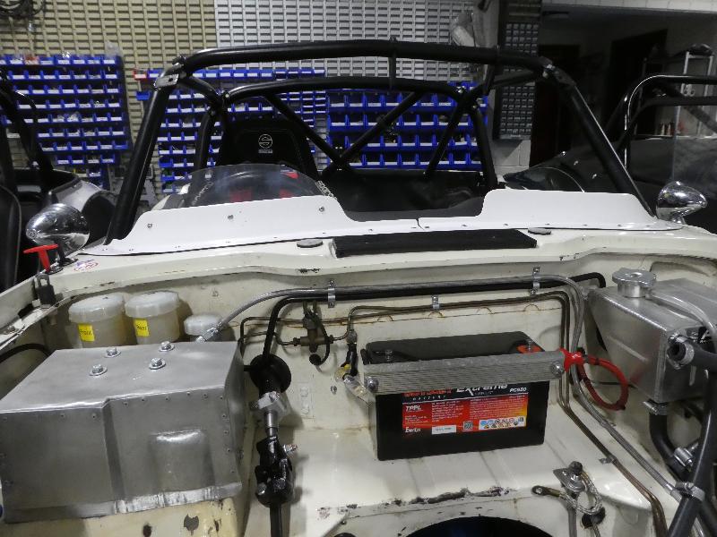 The image shows a TR6 race car from the rear with its engine bay exposed, displaying parts like the battery and wiring. In the background, blue storage bins can be seen. Featured product: Racetorations Wind Deflector - TR4-6.