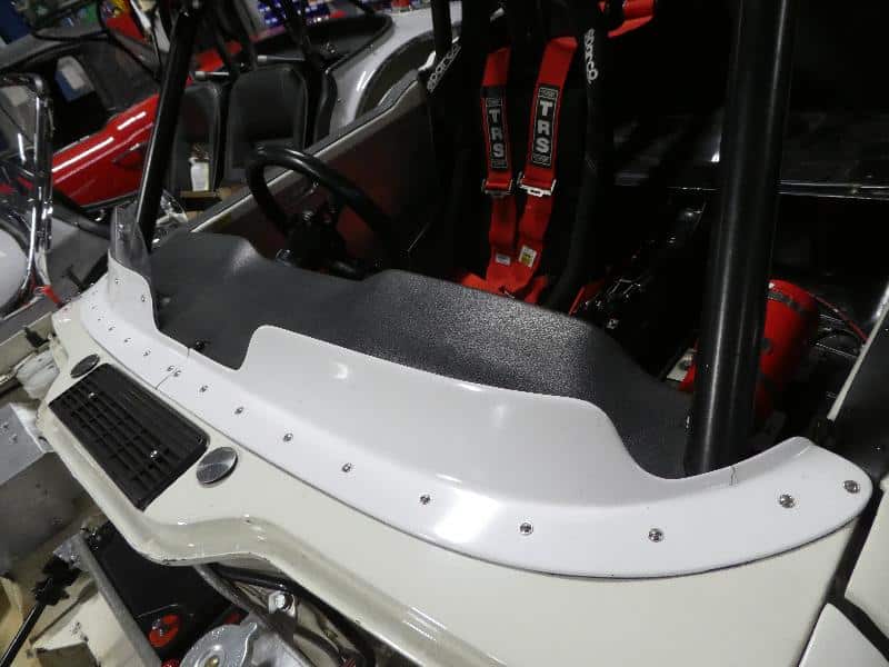 Close-up of a TR4-6 race car cockpit featuring the Racetorations Wind Deflector - TR4-6, black steering wheel, red TRS racing harnesses, white riveted dashboard, and protective roll cage.