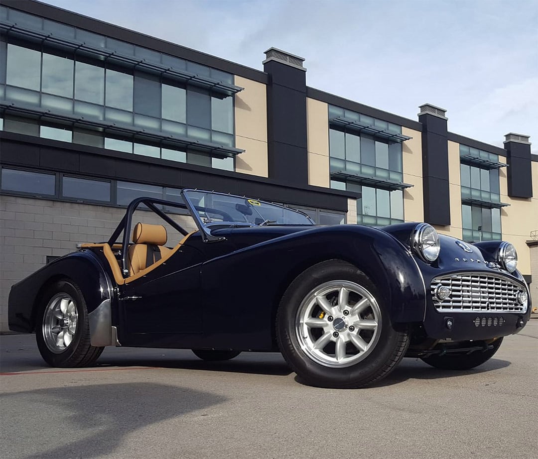 Front three-quarter view of a dark blue classic Triumph TR convertible with tan interior parked outside a commercial building.