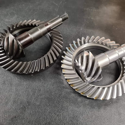 Close-up of two metal bevel gears placed on a dark surface. Each gear set consists of a circular ring gear and a pinion gear, showcasing intricate teeth patterns designed for mechanical transmissions.
